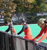 A group of athletes wearing orange uniforms are on the sideline of a sports field, resting against a fence. Behind them, a large digital scoreboard displays team names and scores. The field is bright green, and several banners with sponsor logos and championship wins are visible. The athletes appear to be taking a break, possibly before or during a field hockey game.