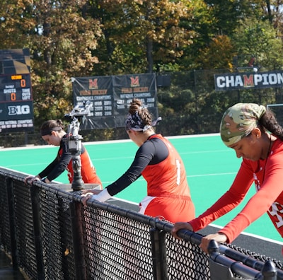 A group of athletes wearing orange uniforms are on the sideline of a sports field, resting against a fence. Behind them, a large digital scoreboard displays team names and scores. The field is bright green, and several banners with sponsor logos and championship wins are visible. The athletes appear to be taking a break, possibly before or during a field hockey game.