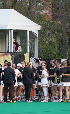 A group of athletes in sports uniforms is gathered on what appears to be a lacrosse field. They are standing in two groups, some wearing white uniforms and some wearing dark uniforms. There is a tent with people inside, possibly officials or commentators, overlooking the field in the background. Extensive greenery and trees are visible behind the tent.