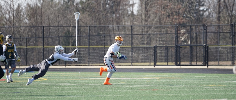 A group of athletes engaged in a sports game on a green field. One player is running forward with possession of the ball, wearing colorful socks and a checkered uniform, while another player in dark clothing dives to intercept or tackle. Other players are present in the background. Leafless trees and a metal fence surround the field.
