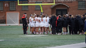 A group of athletes wearing sports uniforms huddles on a grassy field, likely preparing or strategizing for a game. The players are surrounded by additional team members or coaches wearing black jackets. A goalpost and net are visible in the background, along with a brick building.