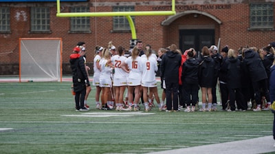 A group of athletes wearing sports uniforms huddles on a grassy field, likely preparing or strategizing for a game. The players are surrounded by additional team members or coaches wearing black jackets. A goalpost and net are visible in the background, along with a brick building.