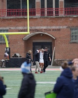 A group of people on a sports field, with some dressed in athletic gear, and others in casual clothing. A brick building labeled 'Gosset Football Team House' is in the background. One of the athletes appears to be aided by others, suggesting a possible injury.