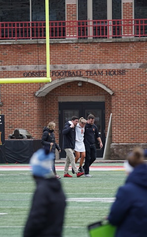 A group of people on a sports field, with some dressed in athletic gear, and others in casual clothing. A brick building labeled 'Gosset Football Team House' is in the background. One of the athletes appears to be aided by others, suggesting a possible injury.