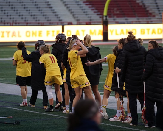 A group of athletes wearing yellow sports uniforms are standing on a sports field. Some are holding lacrosse sticks, and they appear to be interacting casually. Several people are dressed in black coats. In the background, there is a bright sign with text and parts of a stadium are visible.