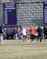 A sports team is practicing or playing on a grassy field in front of a large stone wall with banners displaying men's soccer achievements. People are wearing sports attire, and some are engaged in conversation or coaching.