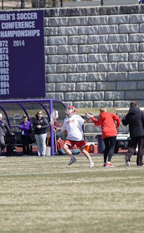 A sports team is practicing or playing on a grassy field in front of a large stone wall with banners displaying men's soccer achievements. People are wearing sports attire, and some are engaged in conversation or coaching.