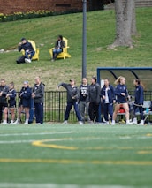 A group of female athletes, some in sports uniforms and others in jackets, are gathered along the sideline of a sports field. They seem to be in a team environment, possibly preparing for or observing a game. In the background, there are a few people sitting on yellow chairs on a grassy incline, and a building with a brick facade.