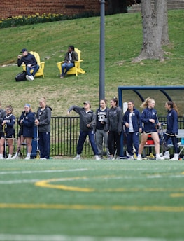 A group of female athletes, some in sports uniforms and others in jackets, are gathered along the sideline of a sports field. They seem to be in a team environment, possibly preparing for or observing a game. In the background, there are a few people sitting on yellow chairs on a grassy incline, and a building with a brick facade.