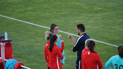 A group of people wearing sports attire are gathered on a grassy field. Some individuals are dressed in red jackets, while others are in turquoise and navy blue. A coach appears to be giving instructions. Sports equipment, such as water bottles and a bench with towels, can be seen nearby.