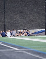 A group of people, likely a sports team, is sitting together on a field. They are wearing matching athletic uniforms, suggesting team unity, and seem to be engaged in a discussion or meeting. In the background, there is sports equipment and a high fence.