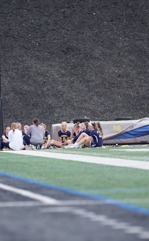A group of people, likely a sports team, is sitting together on a field. They are wearing matching athletic uniforms, suggesting team unity, and seem to be engaged in a discussion or meeting. In the background, there is sports equipment and a high fence.
