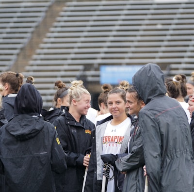 A group of people, likely athletes, are gathered together on a sports field. They are wearing black jackets with some individuals dressed in white sports uniforms underneath. A few people have their hair tied up in buns, and most are engaged in conversation. The background shows tiered stadium seats, suggesting the setting is an outdoor sports venue.