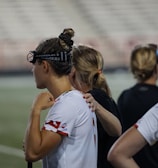 Several people wearing sports attire and protective eyewear are gathered on a sports field. They appear to be engaged in a discussion or briefing, possibly during a break from a game or practice. The background shows a blurred stadium seating, indicating an outdoor setting.
