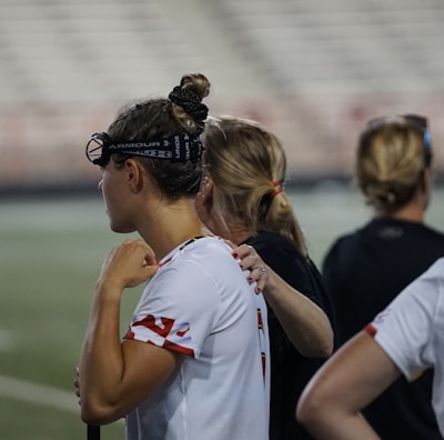 Several people wearing sports attire and protective eyewear are gathered on a sports field. They appear to be engaged in a discussion or briefing, possibly during a break from a game or practice. The background shows a blurred stadium seating, indicating an outdoor setting.