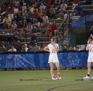 Athletes wearing white sports uniforms stand on a field with lacrosse sticks on the ground. They appear to be part of a lacrosse game, and a large audience is visible in the background seated on bleachers. The environment suggests a competitive sports event.