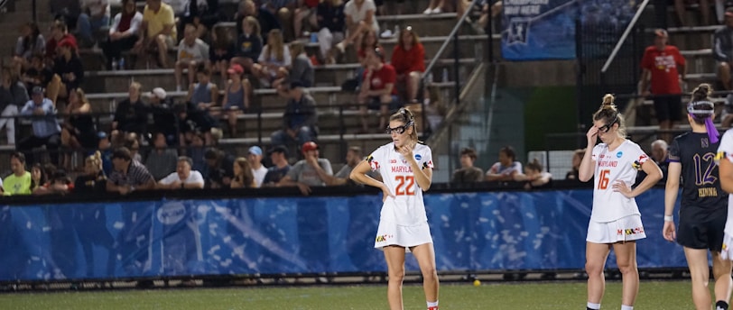 Athletes wearing white sports uniforms stand on a field with lacrosse sticks on the ground. They appear to be part of a lacrosse game, and a large audience is visible in the background seated on bleachers. The environment suggests a competitive sports event.