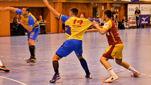 A dynamic indoor sports scene with players in motion on a court. Two athletes in blue and yellow uniforms are engaged in an intense handball game, with one player gripping the other's jersey. The background shows other players, several seated people, and advertisements on the walls.