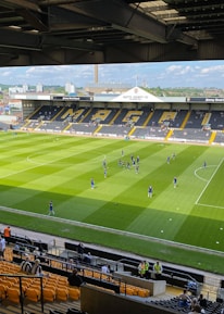A sports stadium with a well-maintained grass field is depicted. Players are spread across the field, engaging in warm-up activities. The stands are partially filled with spectators, and the stadium is surrounded by urban buildings under a partly cloudy sky.