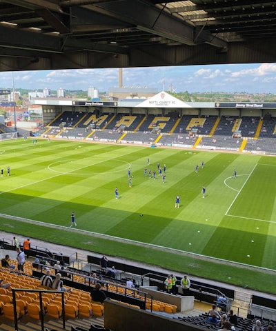 A sports stadium with a well-maintained grass field is depicted. Players are spread across the field, engaging in warm-up activities. The stands are partially filled with spectators, and the stadium is surrounded by urban buildings under a partly cloudy sky.